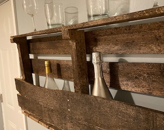May include: A rustic wooden wine rack with a dark brown stain. The rack has two shelves and a bottle holder on the bottom. There are four glasses on the top shelf and two bottles in the bottle holder.