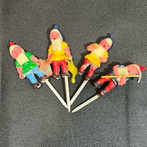 Peut inclure: Ensemble de quatre figurines de gnomes en plastique coloré pour gâteaux. Chaque gnome porte un chapeau rouge et tient un accessoire différent. Les gnomes sont sur des bâtonnets en plastique blanc.