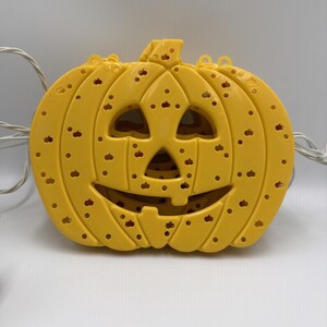 May include: A yellow plastic jack-o'-lantern light cover featuring a carved face with triangular eyes, a triangular nose, and a wide smiling mouth. The surface is covered in small, scattered circular cutouts and includes a white electrical cord.