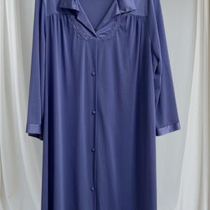 Könnte beinhalten: Ein langer, periwinkelfarbener Morgenmantel mit Kragen und Knopfleiste vorne. Der Morgenmantel hat eine dekorative Stickerei am Ausschnitt und einen Satintrim an den Manschetten und am Kragen. Der Morgenmantel hängt an einem Holzbügel.