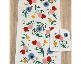 May include: White bath towel and washcloth set with a floral pattern. The design features red, pink, blue, and yellow flowers with green leaves. The towel is rectangular, and the washcloth is square.