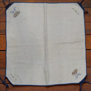 Puede incluir: Un paño cuadrado de lino blanquecino con un borde azul y lazos en las esquinas. Las esquinas presentan diseños florales bordados en tonos rosa, azul y amarillo. El paño se muestra sobre una superficie de madera.