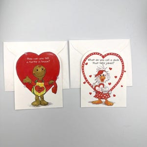 May include: Two heart-shaped greeting cards with white envelopes. The left card features a turtle and the text, How can you tell a turtle is brave? The right card features a duck and the text, What do you call a duck that tells jokes?
