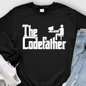 May include: Black sweatshirt with the text "The Codefather" in white, featuring a stylized computer user icon. The sweatshirt is paired with light-wash denim jeans and white sneakers, creating a casual ensemble.