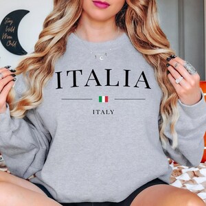 May include: A light gray sweatshirt with the word "ITALIA" printed in black. Below the word is a horizontal line, the Italian flag, and the word "ITALY". The sweatshirt is a crew neck style.