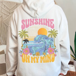 May include: A white hoodie with a graphic print of a sunset, palm trees, a wave, and flowers. The text 'Sunshine on my mind' is printed in pink and yellow letters.