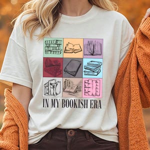 K&ouml;nnte beinhalten: Cremefarbenes T-Shirt mit einem bunten Raster von Buchillustrationen. Der Schriftzug "IN MY BOOKISH ERA" ist darunter gedruckt. Die Person tr&auml;gt eine rostorangefarbene Strickjacke.