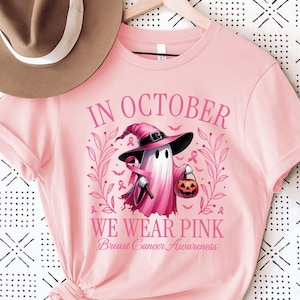 May include: Pink t-shirt with the text "IN OCTOBER WE WEAR PINK Breast Cancer Awareness". The shirt features a ghost wearing a witch hat and holding a pumpkin. A brown hat hangs above the shirt.