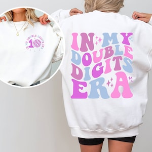 May include: White sweatshirt with the text "IN MY DOUBLE DIGITS ERA" in pink, blue, and purple. The front of the sweatshirt has the number "10" with a disco ball graphic and the text "IN MY DOUBLE DIGITS ERA".