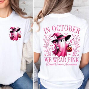 May include: White t-shirt featuring a pink ghost wearing a witch's hat and carrying a pumpkin. The back of the shirt reads "In October We Wear Pink Breast Cancer Awareness".
