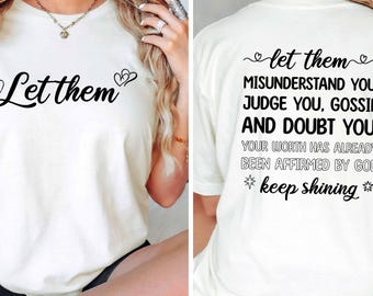 May include: Cream-colored t-shirt with black text on the front that says "Let them" with a heart symbol. The back of the shirt has a longer message in black text, including "keep shining".