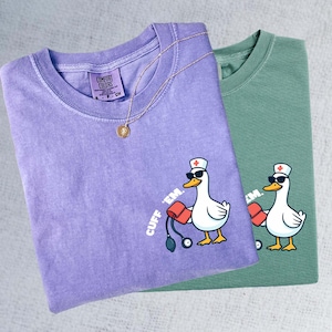 K&ouml;nnte beinhalten: Zwei T-Shirts, eines in Lavendel und eines in Salbeigr&uuml;n, zeigen eine Cartoon-Ente mit Sonnenbrille und Krankenschwesterm&uuml;tze, die eine Blutdruckmanschette h&auml;lt. Der Text "CUFF 'EM." ist auf die Shirts gedruckt.