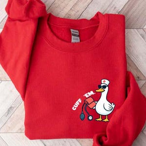 May include: A red sweatshirt with a crew neck. The sweatshirt features a graphic of a white duck wearing sunglasses and a nurse's hat, holding a blood pressure cuff. The text "CUFF 'EM" is printed on the left side.