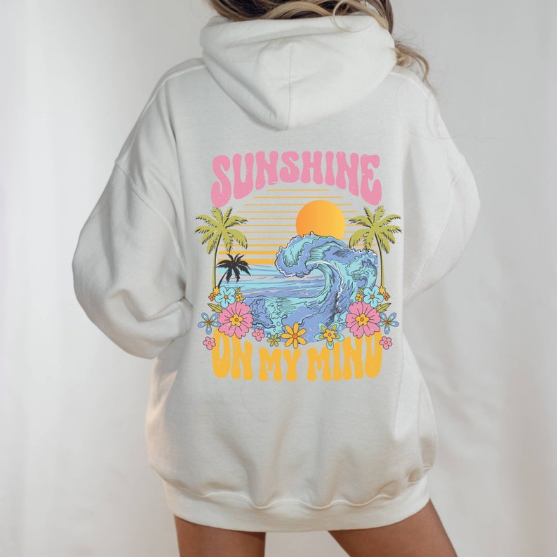 Puede incluir: Sudadera con capucha de color crema con un gr&aacute;fico en la espalda que presenta una puesta de sol, una ola del oc&eacute;ano, palmeras y flores. El texto "SUNSHINE ON MY MIND" est&aacute; en rosa y amarillo. La sudadera tiene capucha y mangas largas.