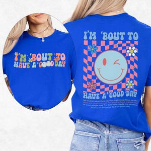 May include: A royal blue t-shirt with the text "I'M 'BOUT TO HAVE A GOOD DAY" in a retro font. The back of the shirt features a smiley face and a checkered pattern with flowers. The shirt has short sleeves.