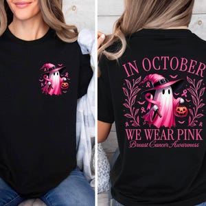 May include: Black t-shirt featuring a pink ghost wearing a witch's hat and holding a pumpkin. The text "In October We Wear Pink Breast Cancer Awareness" is printed on the shirt.