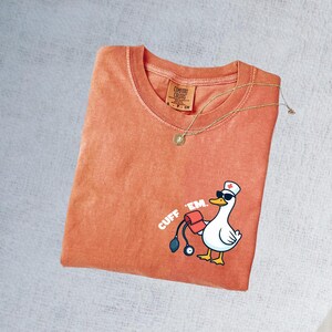 K&ouml;nnte beinhalten: Orangefarbenes Comfort Colors T-Shirt mit einer Cartoon-Gans, die eine Sonnenbrille und eine Krankenschwesterm&uuml;tze tr&auml;gt und eine Blutdruckmanschette h&auml;lt. Der Text "CUFF 'EM" ist auf das Shirt gedruckt. Eine goldene Halskette mit Anh&auml;nger liegt &uuml;ber dem Shirt.