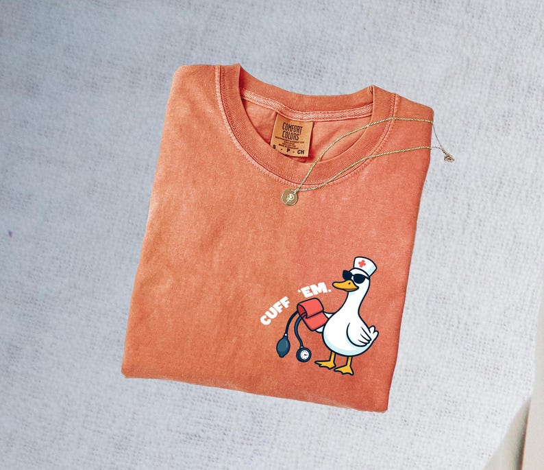 K&ouml;nnte beinhalten: Orangefarbenes Comfort Colors T-Shirt mit einer Cartoon-Gans, die eine Sonnenbrille und eine Krankenschwesterm&uuml;tze tr&auml;gt und eine Blutdruckmanschette h&auml;lt. Der Text "CUFF 'EM" ist auf das Shirt gedruckt. Eine goldene Halskette mit Anh&auml;nger liegt &uuml;ber dem Shirt.