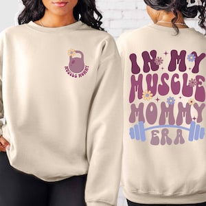 May include: A beige sweatshirt with a pink and white graphic on the front that says "Muscle Mommy" and a pink and white graphic on the back that says "In My Muscle Mommy Era" with a barbell.