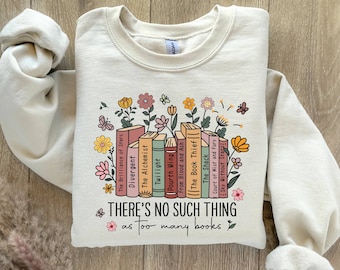 May include: A beige sweatshirt with a graphic of a stack of books with titles like "The Alchemist", "Twilight", and "The Book Thief". The books are surrounded by flowers and butterflies. The text "THERE'S NO SUCH THING as too many books" is printed below the graphic.