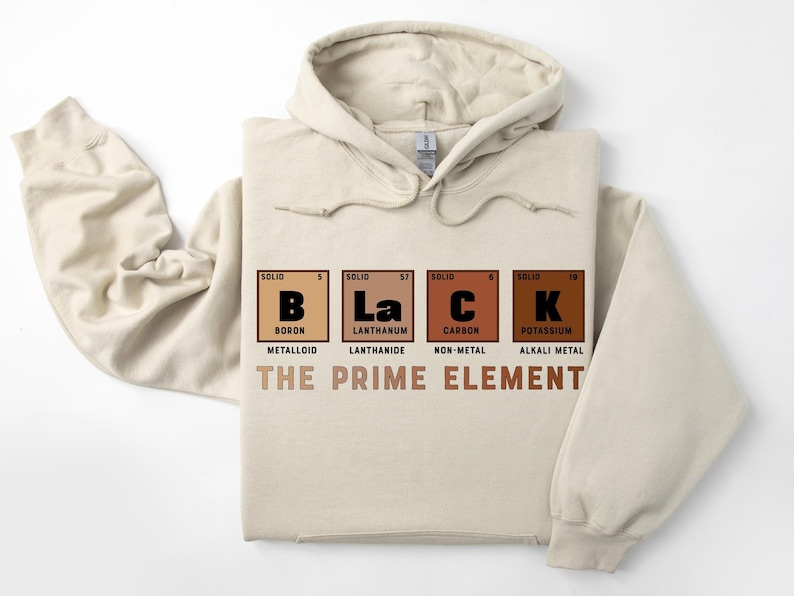 Puede incluir: Una sudadera con capucha beige con un estampado gr&aacute;fico de la tabla peri&oacute;dica de los elementos para el boro, el lantano, el carbono y el potasio, deletreando la palabra "BLACK". El texto "THE PRIME ELEMENT" est&aacute; impreso debajo del gr&aacute;fico.