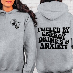 Puede incluir: Sudadera gris con capucha con un gr&aacute;fico negro en la parte delantera de una lata con una mano de esqueleto haciendo el signo de rock and roll. La parte posterior de la sudadera tiene el texto "Fueled by Energy Drinks & Anxiety" en una fuente retro.