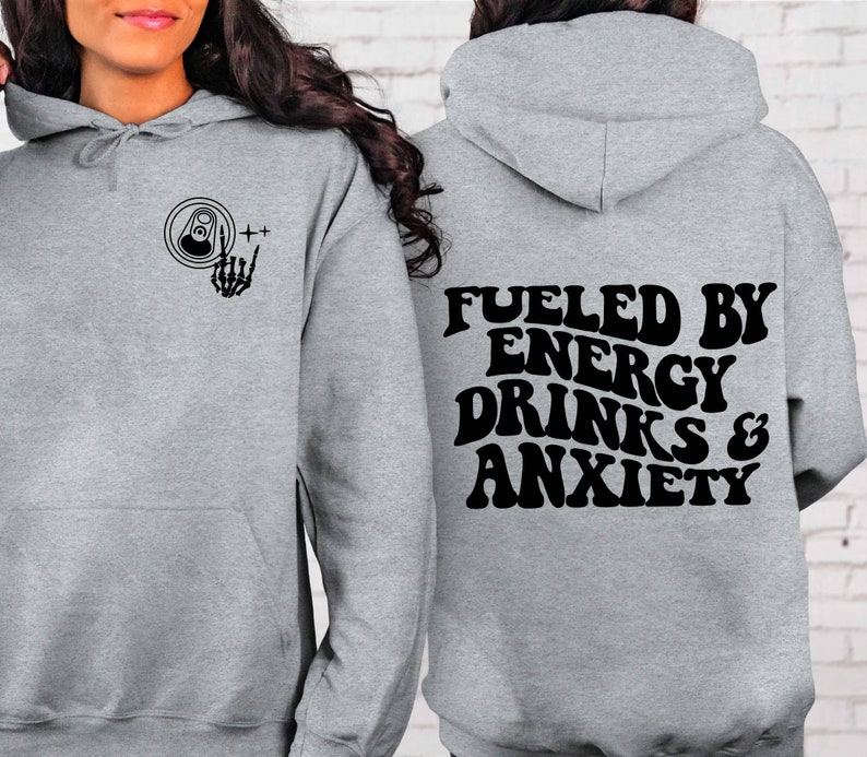 Puede incluir: Sudadera gris con capucha con un gr&aacute;fico negro en la parte delantera de una lata con una mano de esqueleto haciendo el signo de rock and roll. La parte posterior de la sudadera tiene el texto "Fueled by Energy Drinks & Anxiety" en una fuente retro.