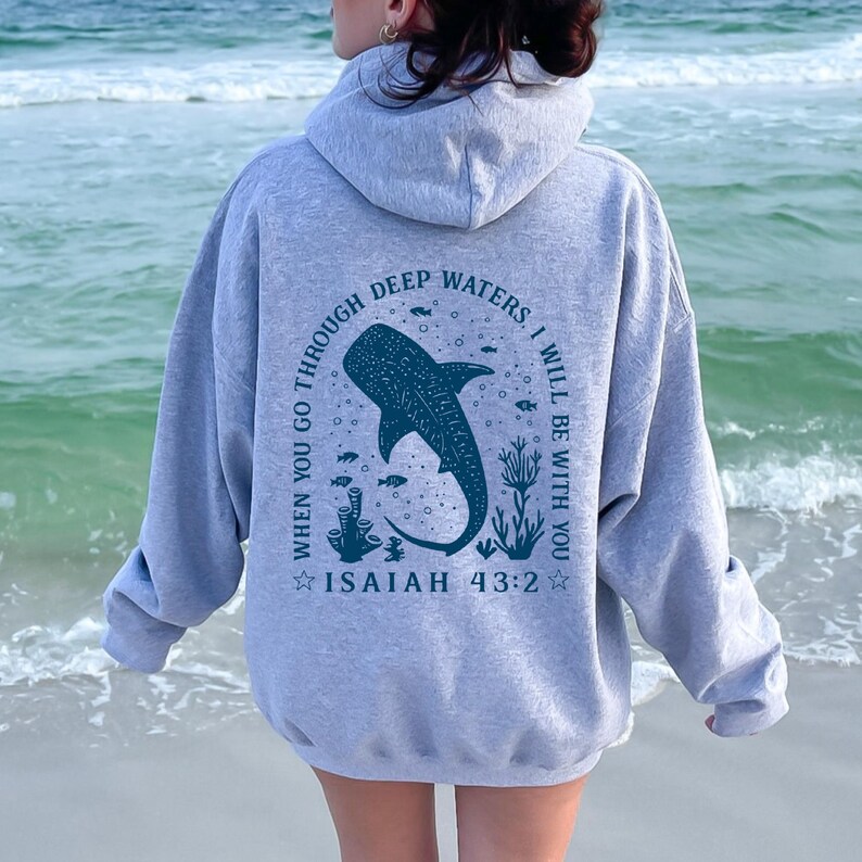 May include: A light gray hoodie with a hood and long sleeves. The back features a blue whale shark illustration with the text "WHEN YOU GO THROUGH DEEP WATERS, I WILL BE WITH YOU. ISAIAH 43:2". The hoodie is made of a soft material.