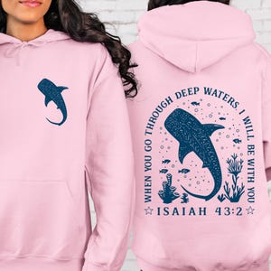 May include: A light pink hoodie featuring a navy blue whale shark graphic on the front and back. The back design includes the text "WHEN YOU GO THROUGH DEEP WATERS, I WILL BE WITH YOU" and "ISAIAH 43:2".