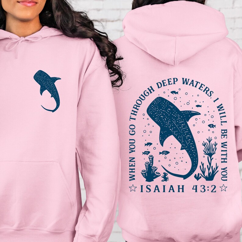 May include: A light pink hoodie featuring a navy blue whale shark graphic on the front and back. The back design includes the text "WHEN YOU GO THROUGH DEEP WATERS, I WILL BE WITH YOU" and "ISAIAH 43:2".