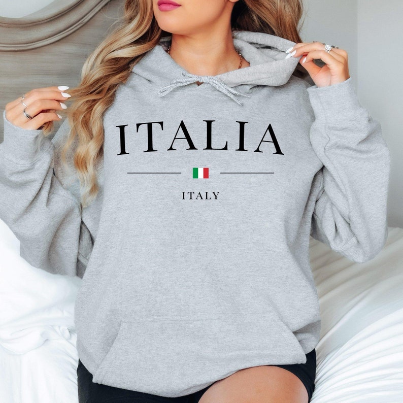 May include: A light gray hoodie with the word "ITALIA" in black capital letters. Below the word is a horizontal line, a small Italian flag, and the word "ITALY". The hoodie has a drawstring hood and a front pocket.