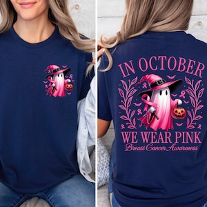 May include: A navy blue t-shirt featuring a pink ghost wearing a witch's hat and holding a pumpkin. The back of the shirt reads "In October We Wear Pink Breast Cancer Awareness".