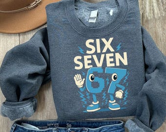 May include: A dark gray sweatshirt with the words "SIX SEVEN" in a cream color. Below the words is the number "67" in blue, designed as cartoon characters. The sweatshirt is paired with blue jeans and a brown hat.