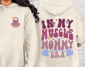 Könnte beinhalten: Ein cremefarbener Hoodie mit einem rosafarbenen Gewichtheber-Design auf der Vorderseite, das "Muscle Mommy" sagt, und einem rosafarbenen, violetten und blauen Design auf der Rückseite, das "In My Muscle Mommy Era" mit einer Langhantel sagt.
