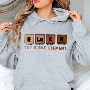 Puede incluir: Sudadera gris con capucha con el texto "BLACK" escrito usando los s&iacute;mbolos qu&iacute;micos de boro, lantano, carbono y potasio. El texto "THE PRIME ELEMENT" est&aacute; impreso debajo de los s&iacute;mbolos qu&iacute;micos.