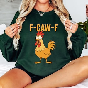 May include: Dark green sweatshirt with the text "F-CAW-F" in orange above a cartoon rooster. The rooster is yellow and orange with a red comb and a wide open beak.