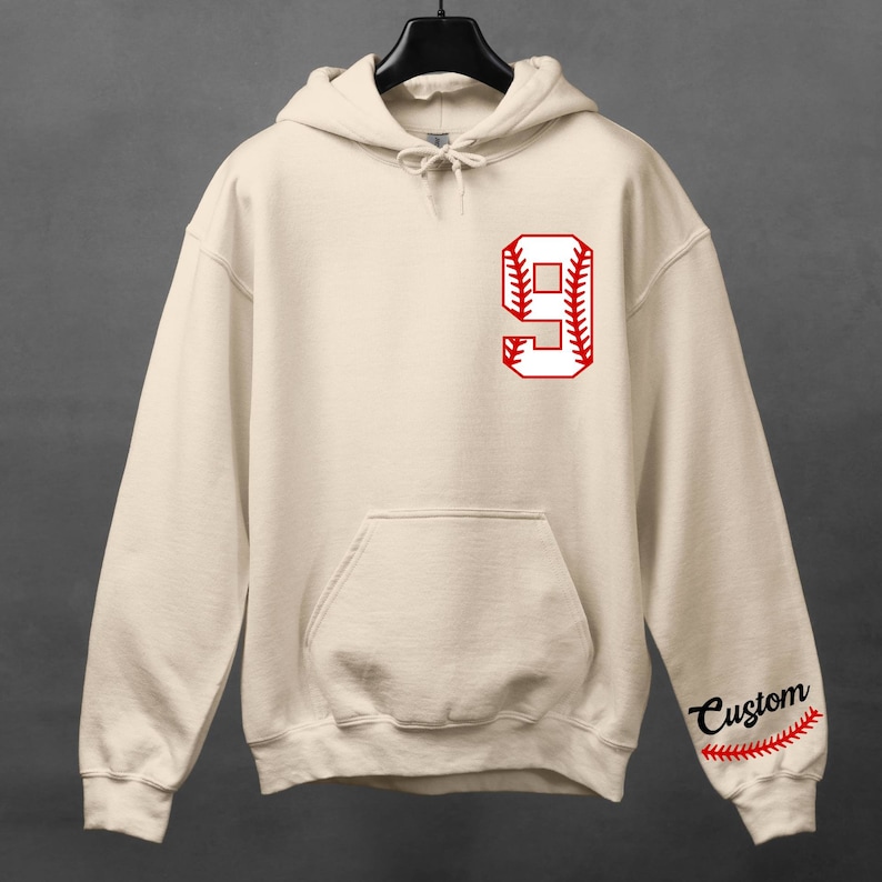 K&ouml;nnte beinhalten: Ein beiger Hoodie mit einer roten Baseball-Nummer 9 auf der Vorderseite und dem Wort "Custom" mit einem roten Baseball-Stich-Design auf dem &Auml;rmel.