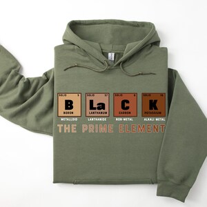 Puede incluir: Una sudadera con capucha verde con la palabra "BLACK" deletreada usando los s&iacute;mbolos qu&iacute;micos del boro, lantano, carbono y potasio. El texto "THE PRIME ELEMENT" est&aacute; impreso debajo de los s&iacute;mbolos qu&iacute;micos.