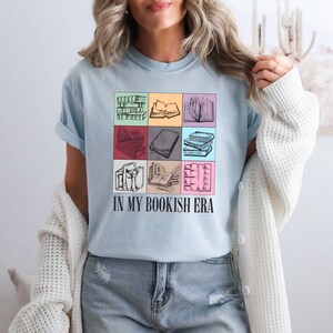 K&ouml;nnte beinhalten: Hellblaues T-Shirt mit einem Raster von Buchillustrationen in verschiedenen Farben. Das Shirt tr&auml;gt den schwarzen Text "IN MY BOOKISH ERA", ein tolles Geschenk f&uuml;r B&uuml;cherw&uuml;rmer.