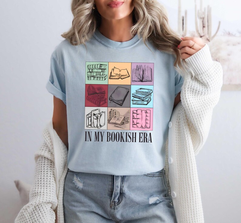 K&ouml;nnte beinhalten: Hellblaues T-Shirt mit einem Raster von Buchillustrationen in verschiedenen Farben. Das Shirt tr&auml;gt den schwarzen Text "IN MY BOOKISH ERA", ein tolles Geschenk f&uuml;r B&uuml;cherw&uuml;rmer.