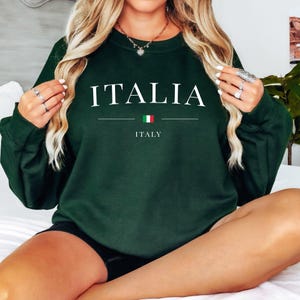 May include: A dark green sweatshirt with the word "ITALIA" in white capital letters. Below the word is a horizontal line with the Italian flag and the word "ITALY" in white. The sweatshirt is a crew neck style.