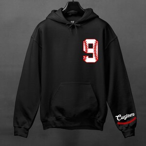 K&ouml;nnte beinhalten: Schwarzer Kapuzenpullover mit einer wei&szlig;en Baseball-Nummer 9 auf der Vorderseite. Die Nummer hat rote N&auml;hte an den R&auml;ndern. Der rechte &Auml;rmel tr&auml;gt das Wort "Custom" in Wei&szlig; mit roten N&auml;hten an den R&auml;ndern.