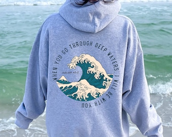 May include: Gray hooded sweatshirt with a blue and white wave graphic on the back. The text "WHEN YOU GO THROUGH DEEP WATERS. I WILL BE WITH YOU" is printed around the wave. The text "ISAIAH 43:2" is printed below the wave.