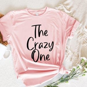 Peut inclure: Un t-shirt rose avec le texte "The Crazy One" imprim&eacute; en noir.