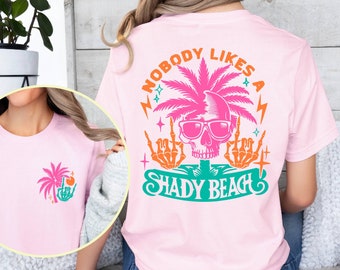 May include: Pink t-shirt with a graphic of a skull wearing sunglasses and a palm tree with the text "Nobody Likes a Shady Beach".