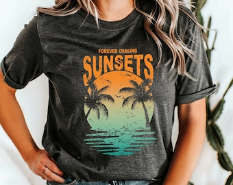 May include: Dark gray t-shirt with a sunset graphic. The graphic features a sun, palm trees, and the words "Forever Chasing Sunsets" in orange. The bottom of the graphic has a teal and blue water design.
