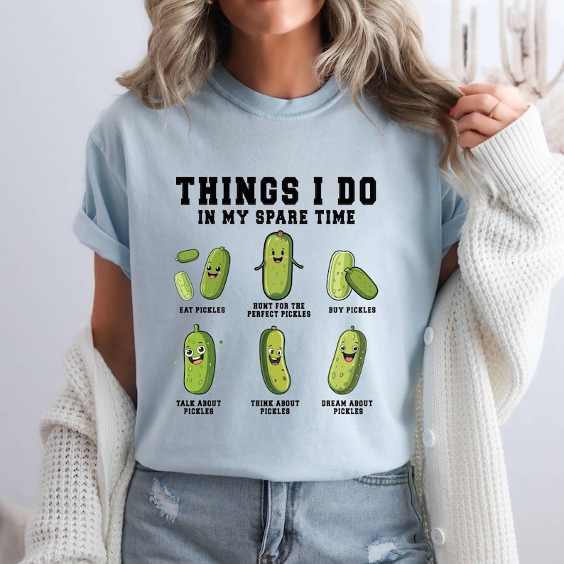 Pu&ograve; includere: Maglietta blu chiaro con una grafica di sottaceti e il testo "THINGS I DO IN MY SPARE TIME". La grafica mostra una serie di sottaceti con diverse espressioni e attivit&agrave;, tra cui mangiare, cacciare, acquistare, parlare, pensare e sognare i sottaceti.