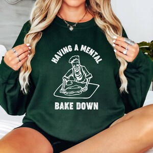 May include: A dark green sweatshirt with a white graphic of a skeleton baking. The text on the sweatshirt reads "Having a mental bake down."