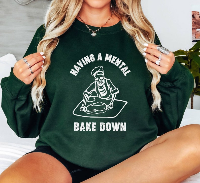 May include: A dark green sweatshirt with a white graphic of a skeleton baking. The text on the sweatshirt reads "Having a mental bake down."