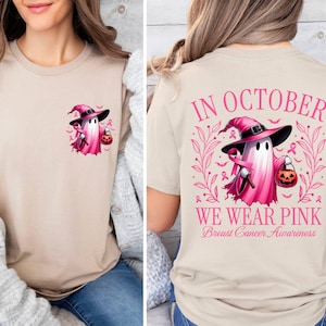 May include: A tan t-shirt with a pink ghost wearing a witch's hat and holding a pumpkin. The back of the shirt says "In October We Wear Pink Breast Cancer Awareness" with a pink ghost wearing a witch's hat and holding a pumpkin.